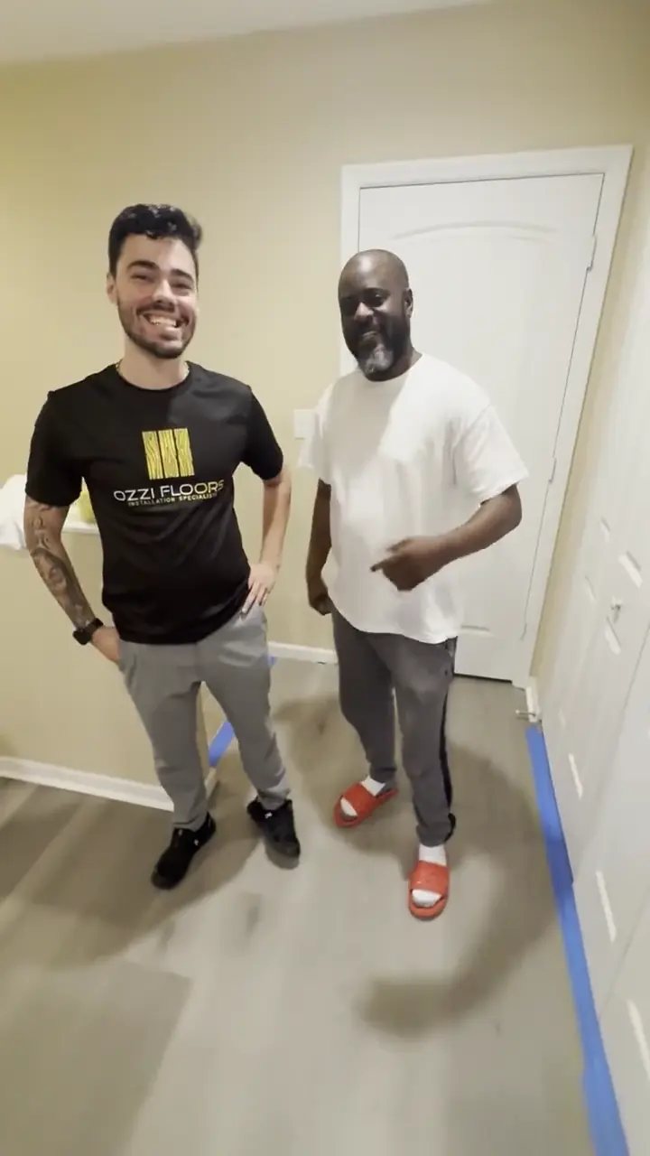 An Ozzifloors team member standing with a customer in a hallway on a newly laid grey plank floor, painter's tape still down along the edges